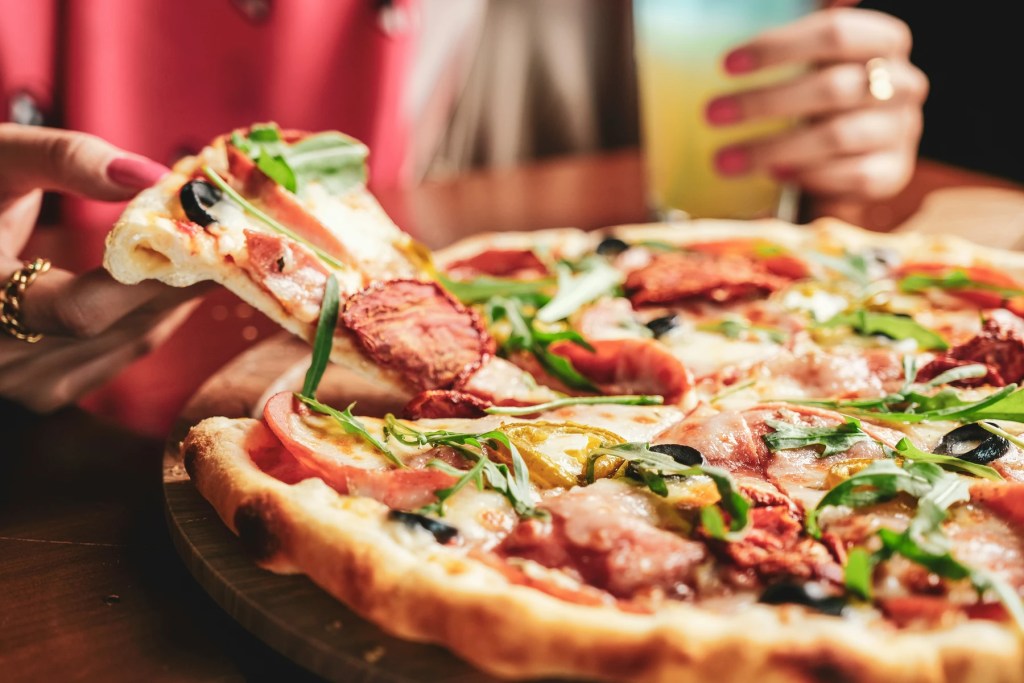 Imagem de uma pizza recheada com queijo, tomates e pimentão. Uma fatia está cortada e sendo retirada por uma mulher, que veste uma camiseta vermelha e usa anéis nas mãos.