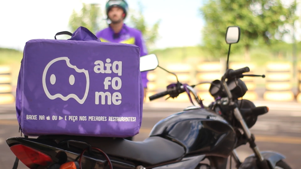 bag aiqfome em destaque com moto e entregador desfocados em segundo plano 